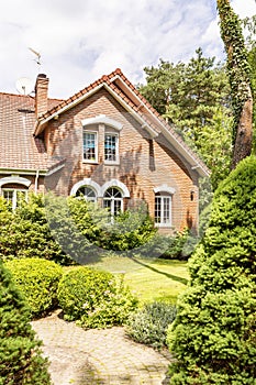 A garden with evergreens, trees, shrubs and grass in the backyard of a red brick luxurious English style house.