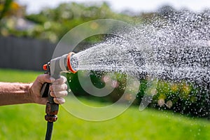 Garden hose with nozzle. Man\'s hand holding spray gun and watering plants, spraying water on grass in backyard