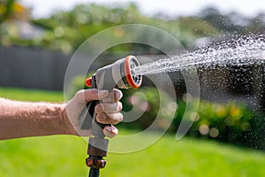 Garden hose with nozzle. Man's hand holding spray gun and watering plants, spraying water on grass in backyard