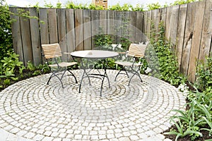 Garden patio with two chairs and round table in front of flowerbed and wooden planks