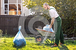The gardener in the green uniform is cleaning the yard.On the grass is a cart with compost and a package of garbage