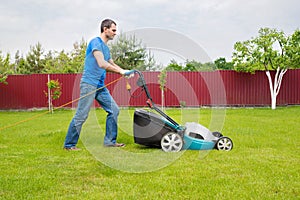 A gardener with a lawn mower is cutting green grass in the garden in the house backyard