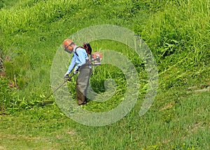 A gardener trims the grass in a downhill field with a weed eater. He wears all the prescribed safety clothes and accessories