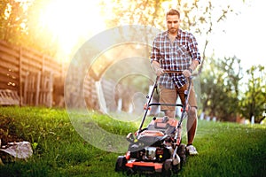 gardener working with lawnmower and cutting grass in backyard during summer sunset