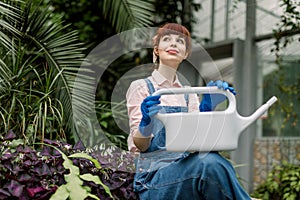 Gardening woman with watering can in greenhouse. Pretty woman agronomist sitting with watering can in modern hothouse
