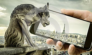 Gargoyle on Cathedral of Notre Dame de Paris looks at Eiffel Tower in mobile or cell phone, Paris, France. Funny photo of old