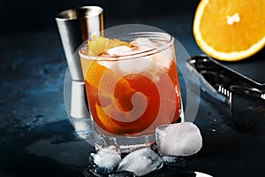 Garibaldi alcoholic cocktail with red bitter, orange juice, zest and ice. Dark blue background, bar tools, selective focus