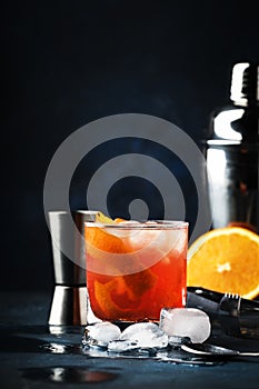 Garibaldi alcoholic cocktail with red bitter, orange juice, zest and ice. Dark blue background, bar tools, selective focus