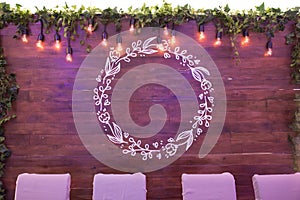 Garland lamps over wooden board fence with green leaves. Empty place for the names of the newlyweds.