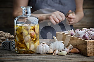 Garlic aromatic flavored oil or infusion bottle and wooden crate of garlic cloves on wooden kitchen table. Woman peels garlic.