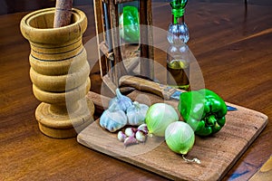 Garlic, onions and green pepper on wooden cutting board with glass of olive oil, wooden mortar and wooden chopping block with kniv