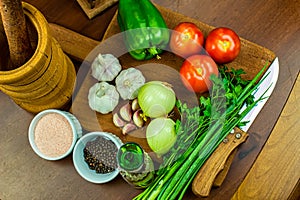 Garlic, onions, green peppers, tomatoes and parsley and chives on wooden cutting board with glass of olive oil, salt and pepper