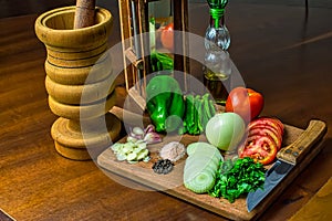 Garlic, onions, green peppers, tomatoes and parsley and chives on wooden cutting board with glass of olive oil, salt and peppers