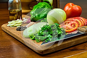 Garlic, onions, green peppers, tomatoes and parsley and chives on wooden cutting board with glass of olive oil, salt and peppers