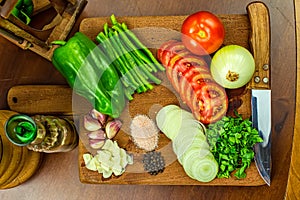 Garlic, onions, green peppers, tomatoes and parsley and chives on wooden cutting board with glass of olive oil, salt and peppers