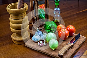 Garlic, onions, green peppers and tomatoes on wooden cutting board with glass of olive oil, wooden mortar and wooden chopping bloc