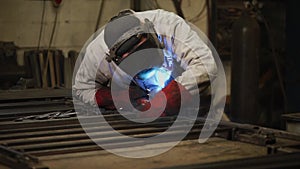 Gas welder is in a shop of plant, leaning over metal beams and welding