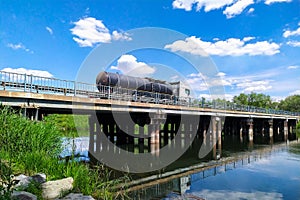 A gasoline or gas tanker truck transports fuel to a gas station through a river bridge on a sunny day