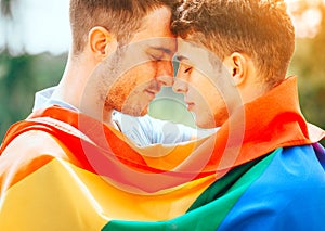 Gay couple with colourful LGBT flag enjoying nature outdoors, kissing and hugging. Young men family in love concept