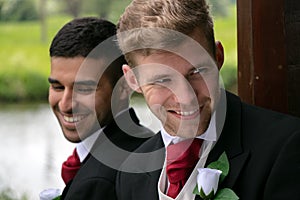 Gay couple of grooms pose for photographs by a lake on their wedding day