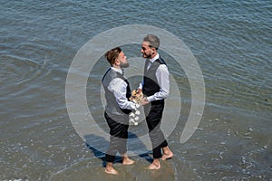Gay grooms walking together on sea beach during Wedding day. Romantic men in sea water. Romantic and sensual gay couple