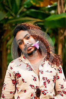 Gay man with curly hair and floral shirt stands in garden, purple flower in mouth. Expresses freedom, LGBT pride, gender