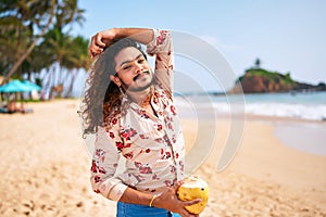 Gay man with stylish hair and floral shirt enjoys fresh coconut on sunny beach. Relaxed male sips nutritious drink