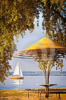 Gazebo surrounded by tree branches on the bay with a sailing yacht on the horizon. Autumn landscape on a sunny day.