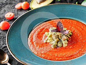 Gazpacho tomato cream soup in plate with avocado and greens over dark background.