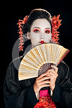 Geisha in black kimono with red flowers in hair holding traditional asian hand fan isolated on black