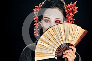 Geisha in black kimono with red flowers in hair holding traditional asian hand fan near face