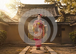 Geisha in a kimono posing on a stone path in front of the gate of