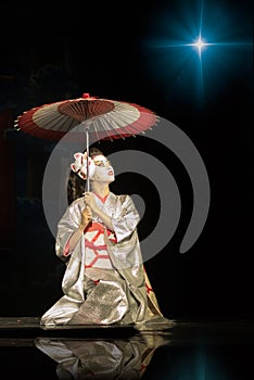 Geisha in traditional kimono with umbrella sitting on the knees in darkness, lighten by the star and looking up.