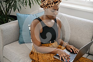 Gen z Black African woman sitting on sofa at home typing on laptop