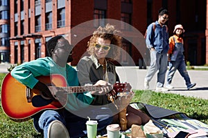 Gen Z young couple enjoying picnic on grass in city and playing guitar
