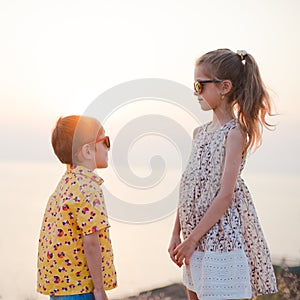 Gender relationship concept of happy little kids boy and girl look at each other through summer sunglasses on sea beach in sunset