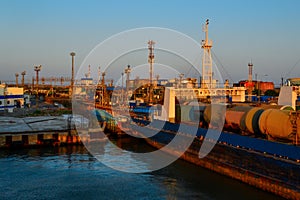 General view of an industrial harbor with moored large ships with masts, barges with cargo, boats in the red light of sunset