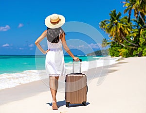 A woman in a white dress and straw hat walks along a pristine beach with a rolling suitcase.