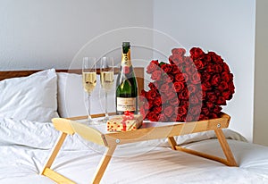 Celebrating Saint Valentine`s Day with bouquet of red roses, glass of champagne and croissants on tray on bed. Romantic surprise