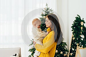 A gentle attentive mother holds a one-year-old child while standing at the Christmas tree at home.