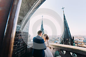 Gentle beautiful bride and groom holding hands embracing face-to-face on the ancient balcony, background cityscape