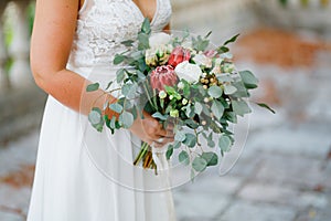 A gentle bride holds in her hands an unusual wedding bouquet of white peonies, roses, proteas and eucalyptus branches