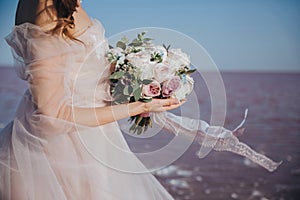 Gentle bride in an ivory wedding dress on the seashore with the bouquet in her hands on the sea background. Back view.