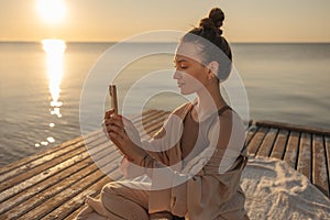 Gentle image of young caucasian girl sitting on wooden pier with gadget in hands on seashore.