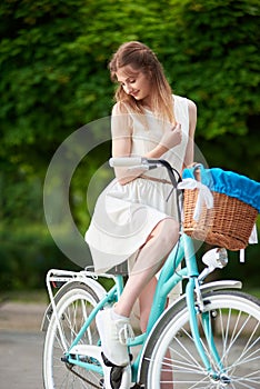 Gentle lady posing while sitting on a retro blue bike with basket in the town park