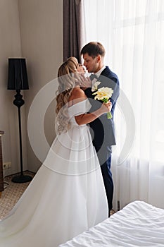 gentle and in love blonde bride and groom kiss by the window.