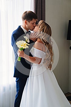 gentle and in love blonde bride and groom kiss by the window.