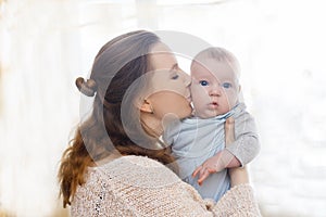 Gentle photo of mother and baby. Mom kisses her son on the cheeks on a light background
