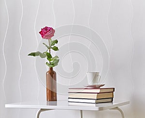 Gentle pink rose in clay bottle, cup of coffee, books on a white coffee table against a light wall.