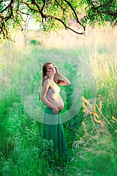 Gentle pregnant in a green skirt and a light blouse stands in the sun in a field